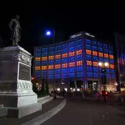 Night view of a city square featuring a statue in the foreground and a modern building illuminated with blue and orange lights in the background. The building displays the AES Indiana logo on its rooftop, and streetlights brighten the plaza.