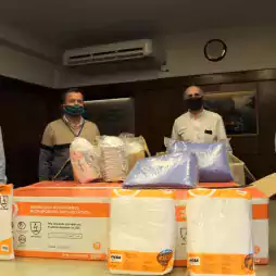 Four men wearing face masks stand behind a table with boxes and packages of disposable protective coveralls and personal protective equipment in an office setting.