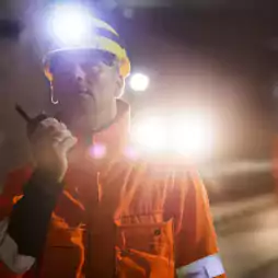 A worker wearing an orange safety jacket and yellow hard hat with a headlamp communicates via walkie-talkie inside a dimly lit underground tunnel. Two other workers in similar attire are visible in the background.