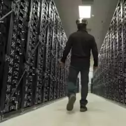 A person wearing a helmet walks down a corridor between two large rows of server racks in a data center, with overhead lighting illuminating the space.