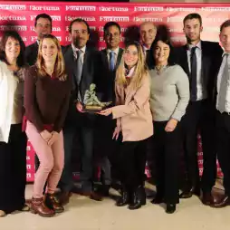 A group of eleven professionally dressed people stand together smiling in front of a red Fortuna backdrop. Two women in the front center hold a trophy or award, suggesting a celebration or recognition event.