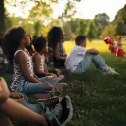 A group of children sit on grass in a park, attentively watching something out of frame. The scene is set in daylight with trees and orange traffic cones visible in the background.